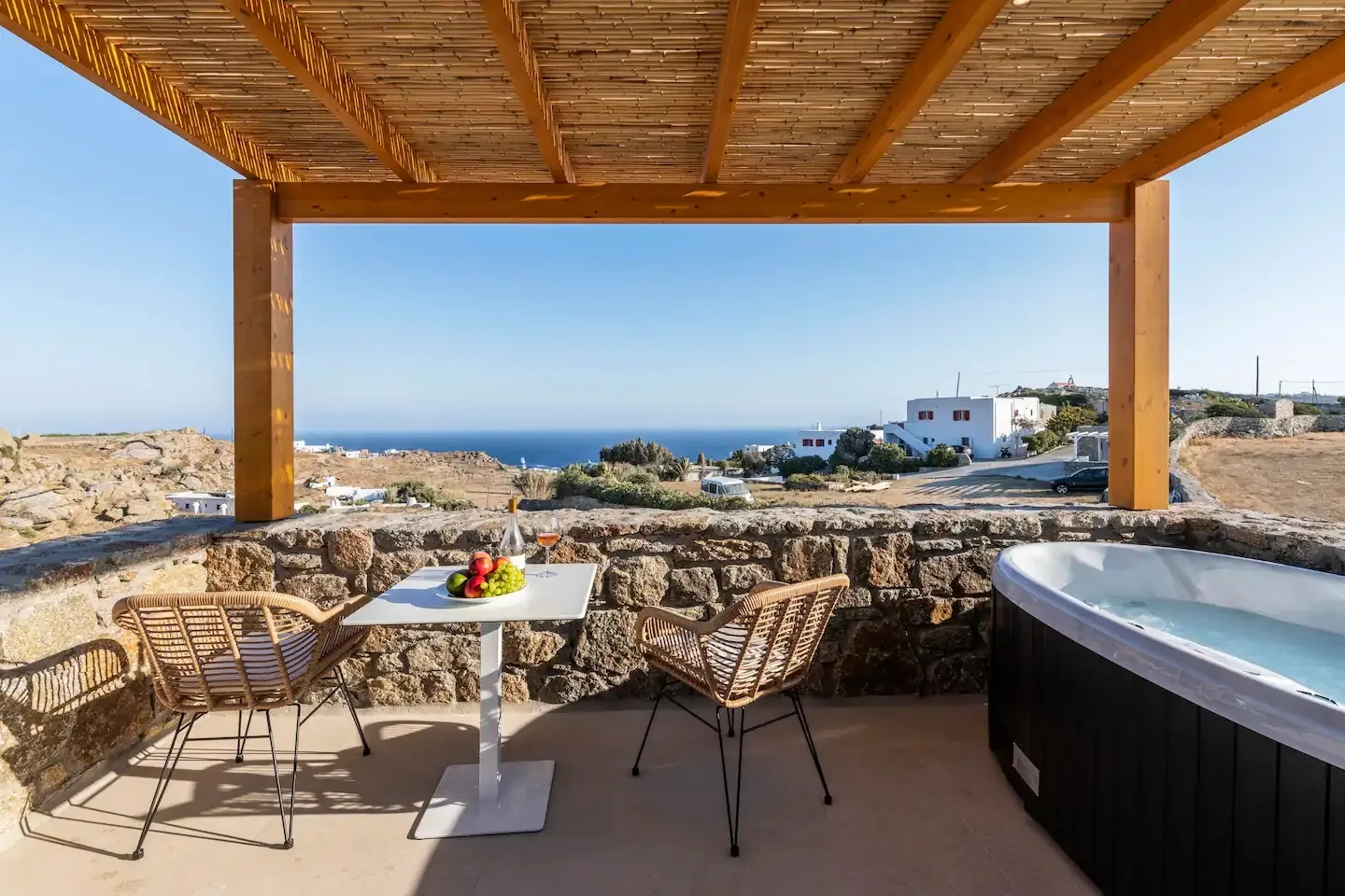 Una zona de comedor exterior a la sombra mira al mar, con mesa y asientos bajo una pérgola de madera.