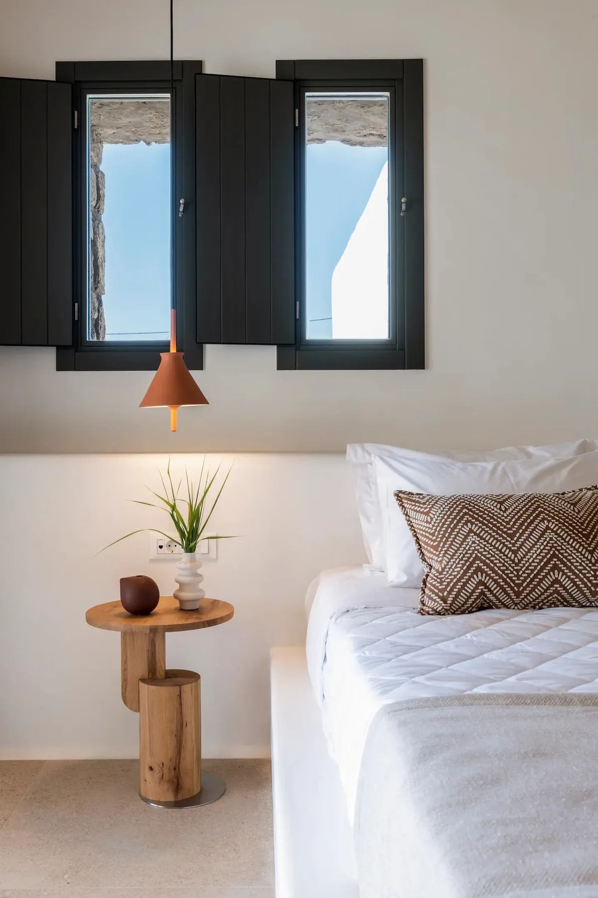 Un dormitorio luminoso con una cama blanca, dos ventanas de marco oscuro y una pequeña mesa de madera.