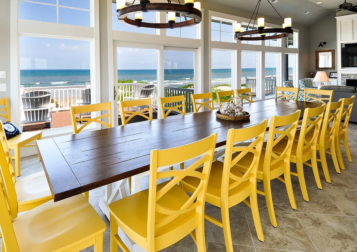 Comedor con una mesa larga y sillas amarillas junto a ventanas con vistas al mar.