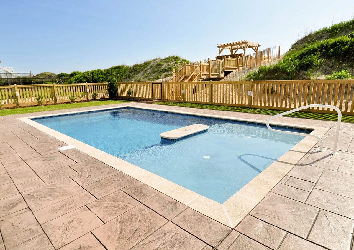Una piscina al aire libre está junto a una terraza pavimentada y una cerca de madera.