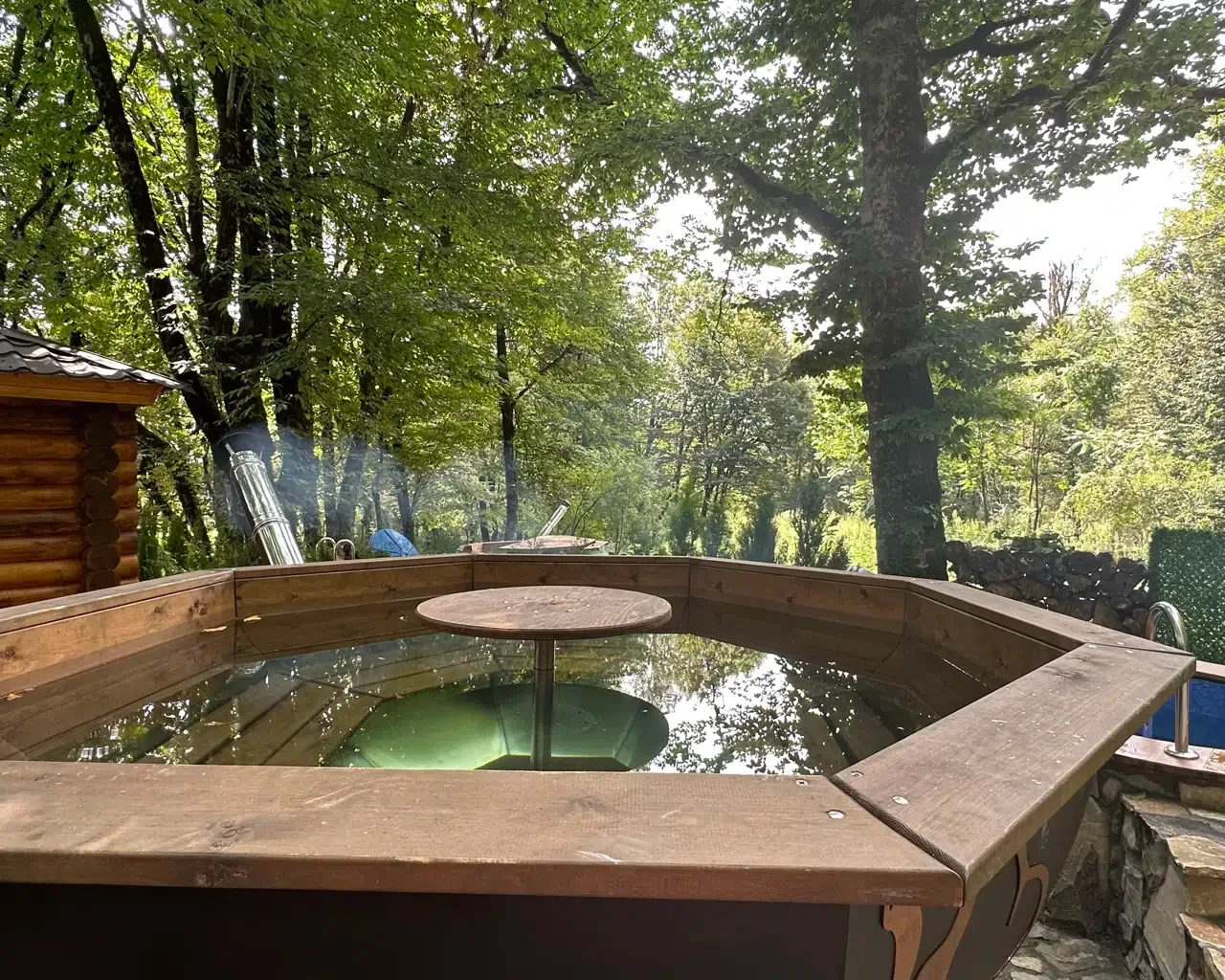 Espacio representativo del hotel: Una bañera de hidromasaje de madera está al aire libre, entre árboles y junto a una cabaña rústica.