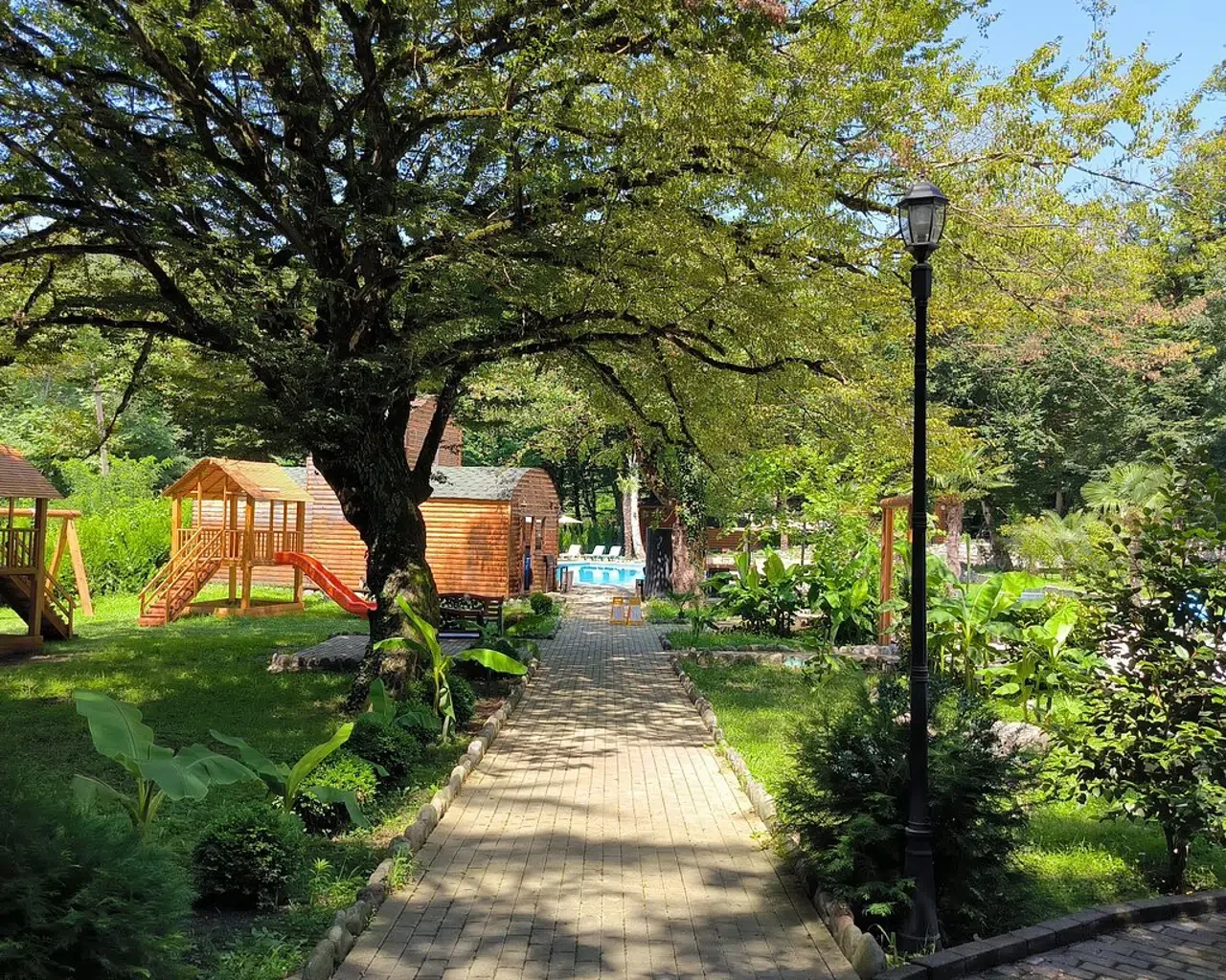Espacio representativo del hotel: Un sendero ajardinado pasa bajo un gran árbol y conduce a juegos infantiles y una piscina.
