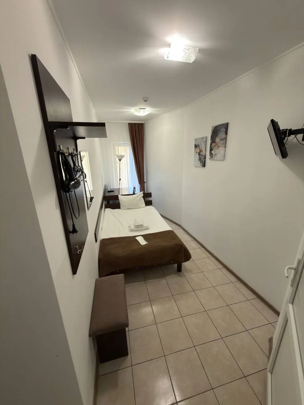 Espacio representativo del hotel: Un dormitorio estrecho con una cama, suelo de baldosas y televisor en la pared.