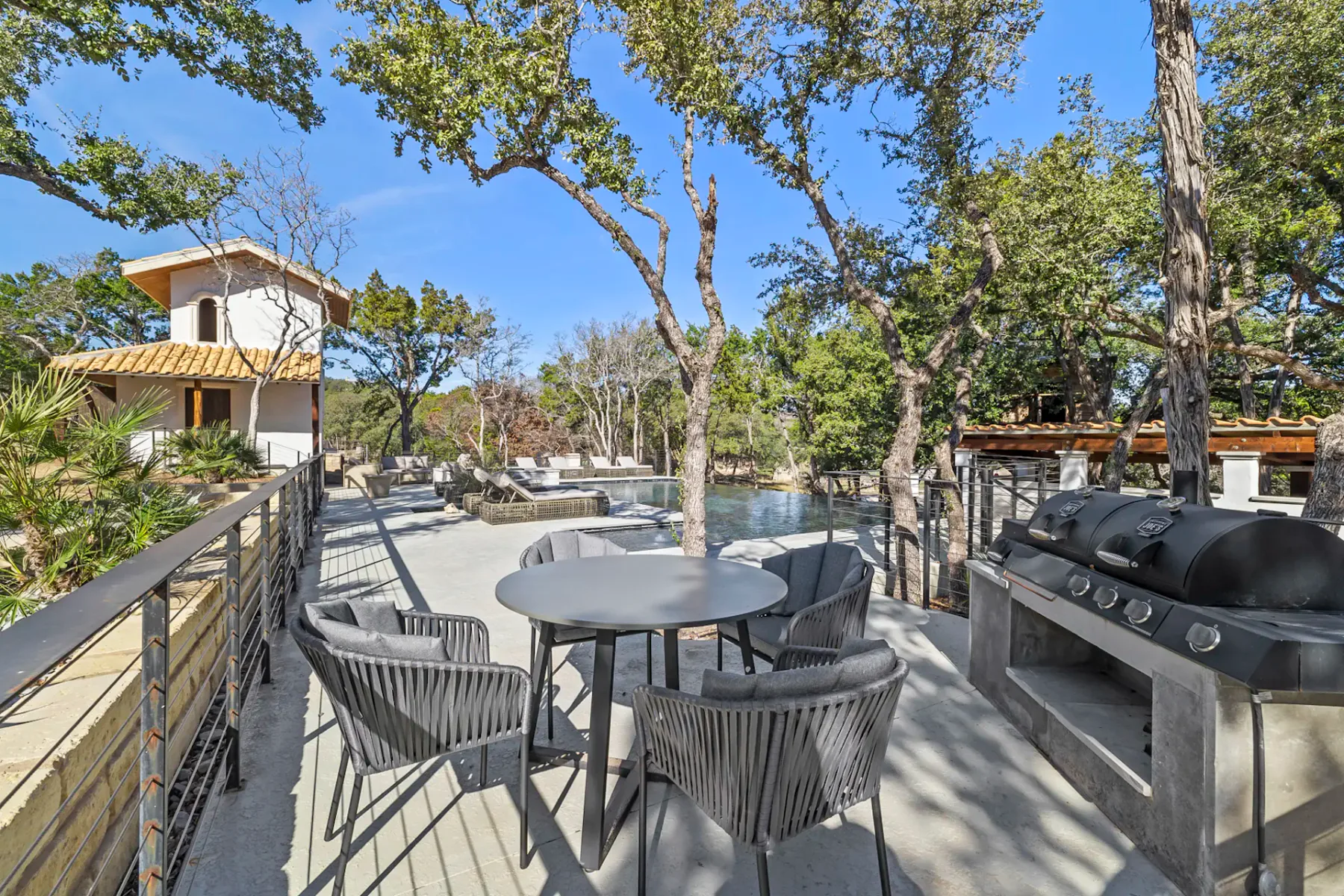 Shared amenity: An outdoor patio has a round dining table, seating, and a grill beneath leafy trees.