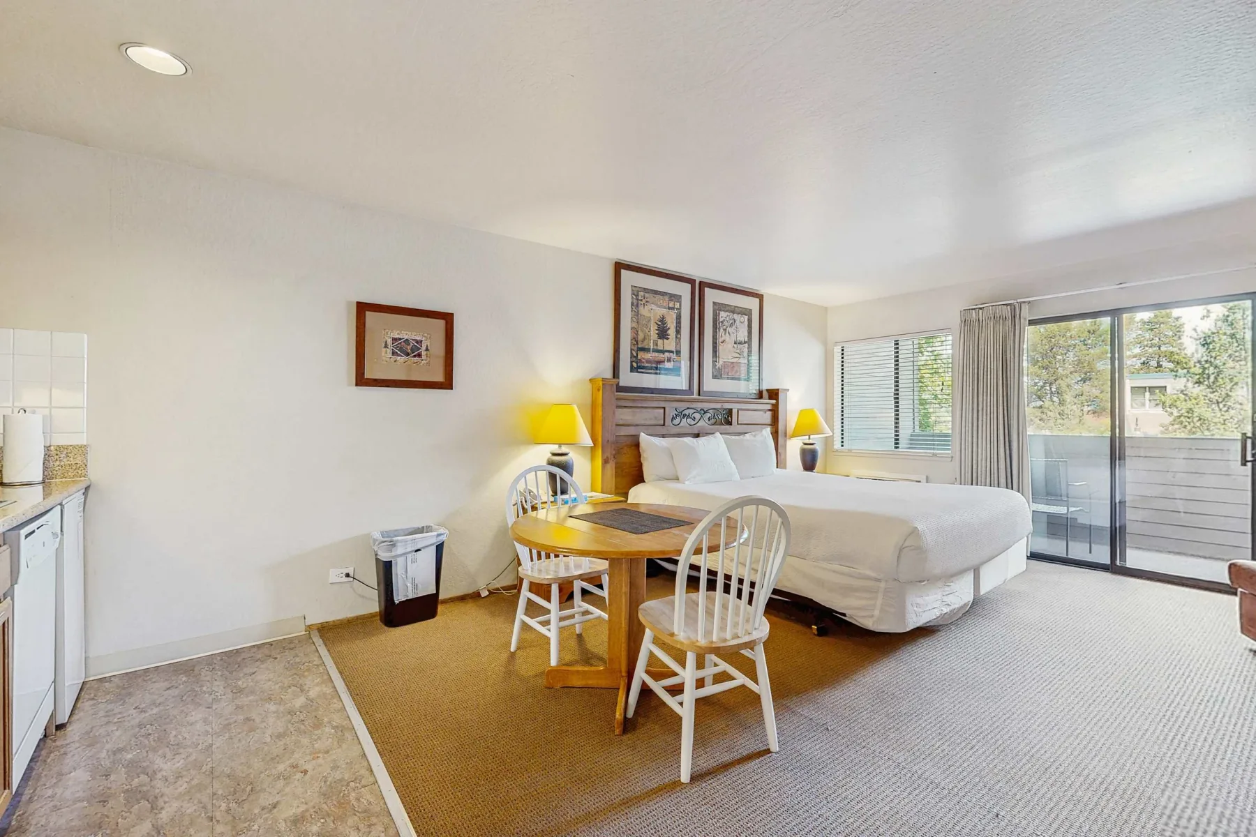 A bright bedroom has a bed, a small dining table, and glass doors opening to a balcony.