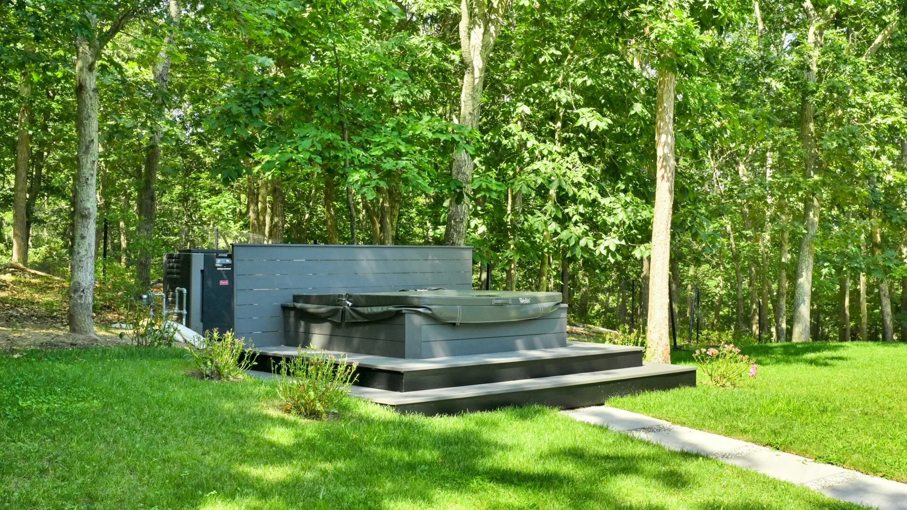 A hot tub sits on a low deck in a grassy clearing surrounded by trees.