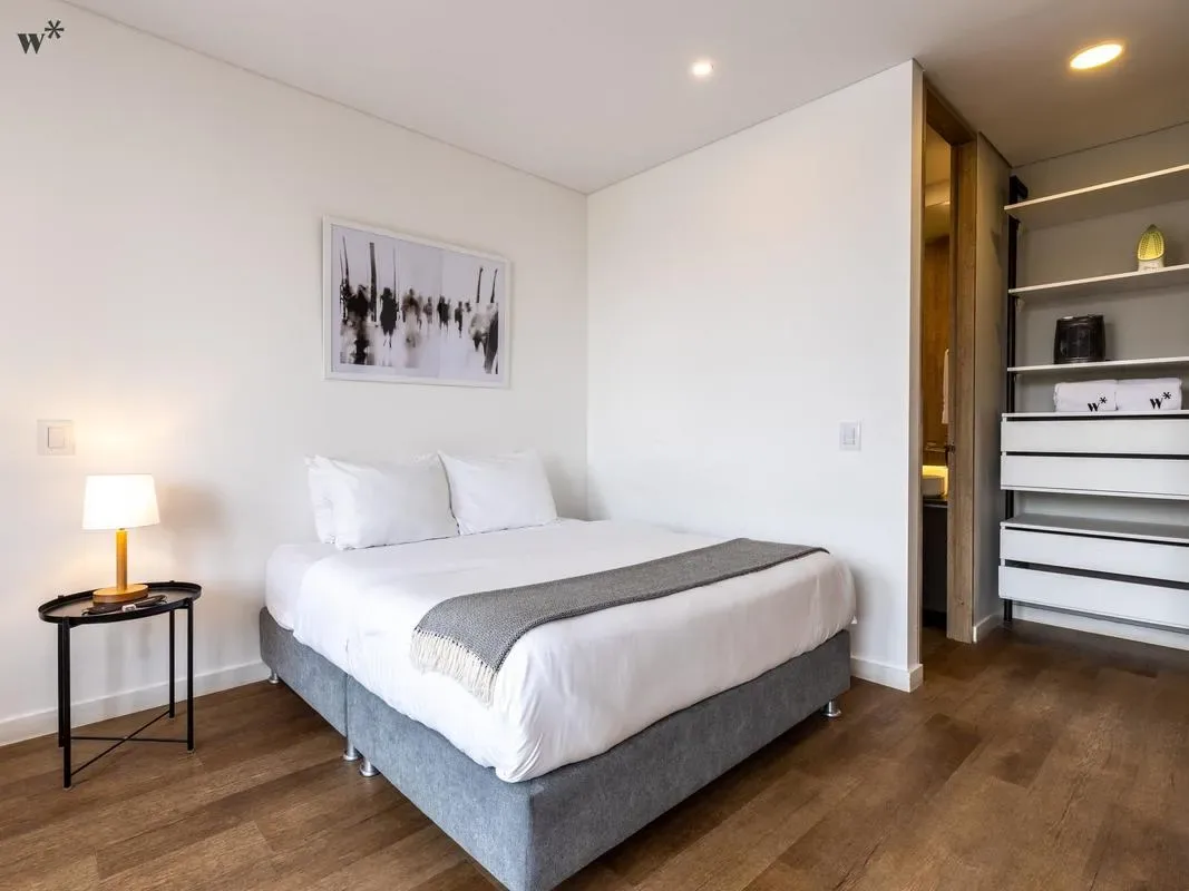 Un dormitorio moderno con una cama blanca, una manta gris, una lámpara de noche y estanterías empotradas.