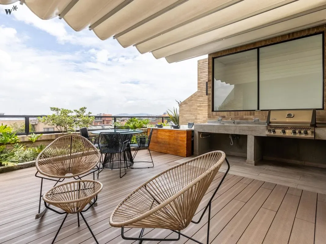 Una terraza en la azotea tiene sillas tejidas, una mesa de comedor, una parrilla empotrada y un toldo retráctil.