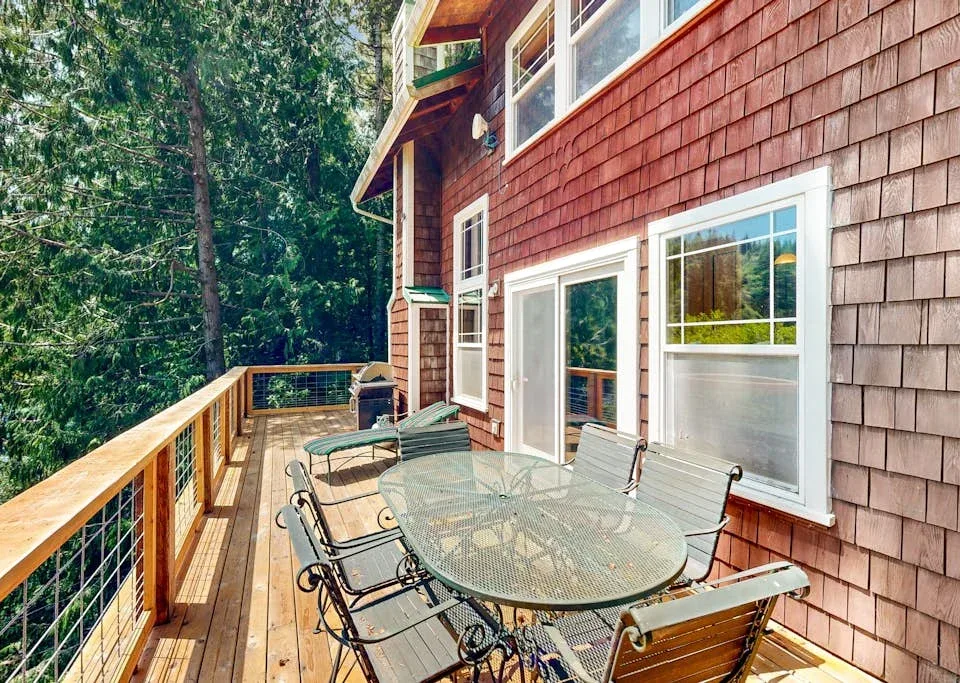 Una terraza exterior con una mesa de vidrio y asientos junto a un paisaje arbolado.
