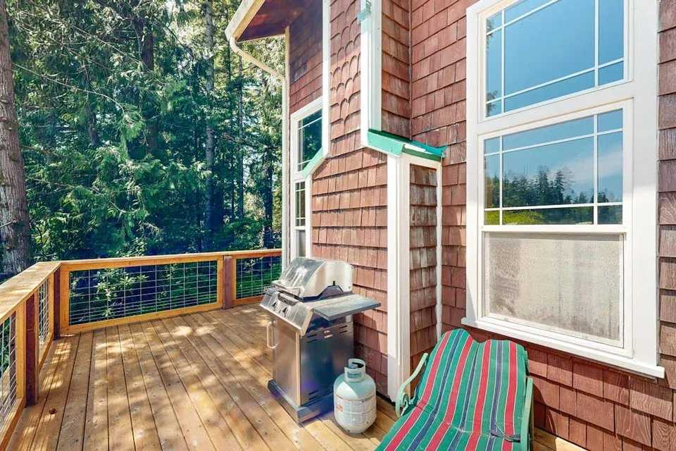 Una terraza de madera junto a una casa tiene una parrilla y asientos al aire libre.