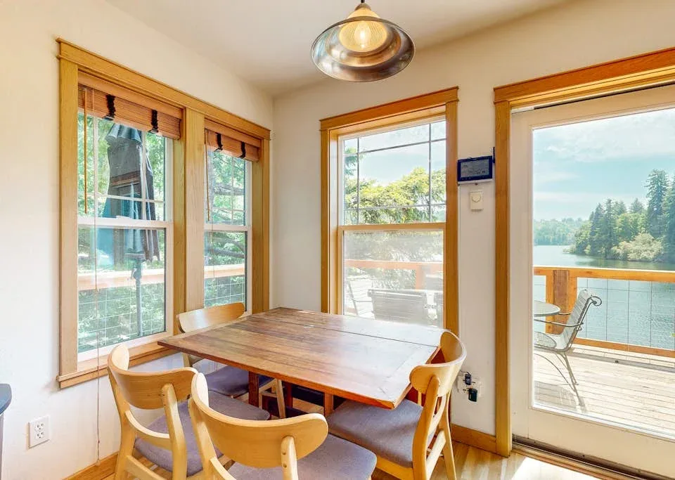 Comedor con una mesa de madera junto a grandes ventanas con vistas a una terraza y al agua.