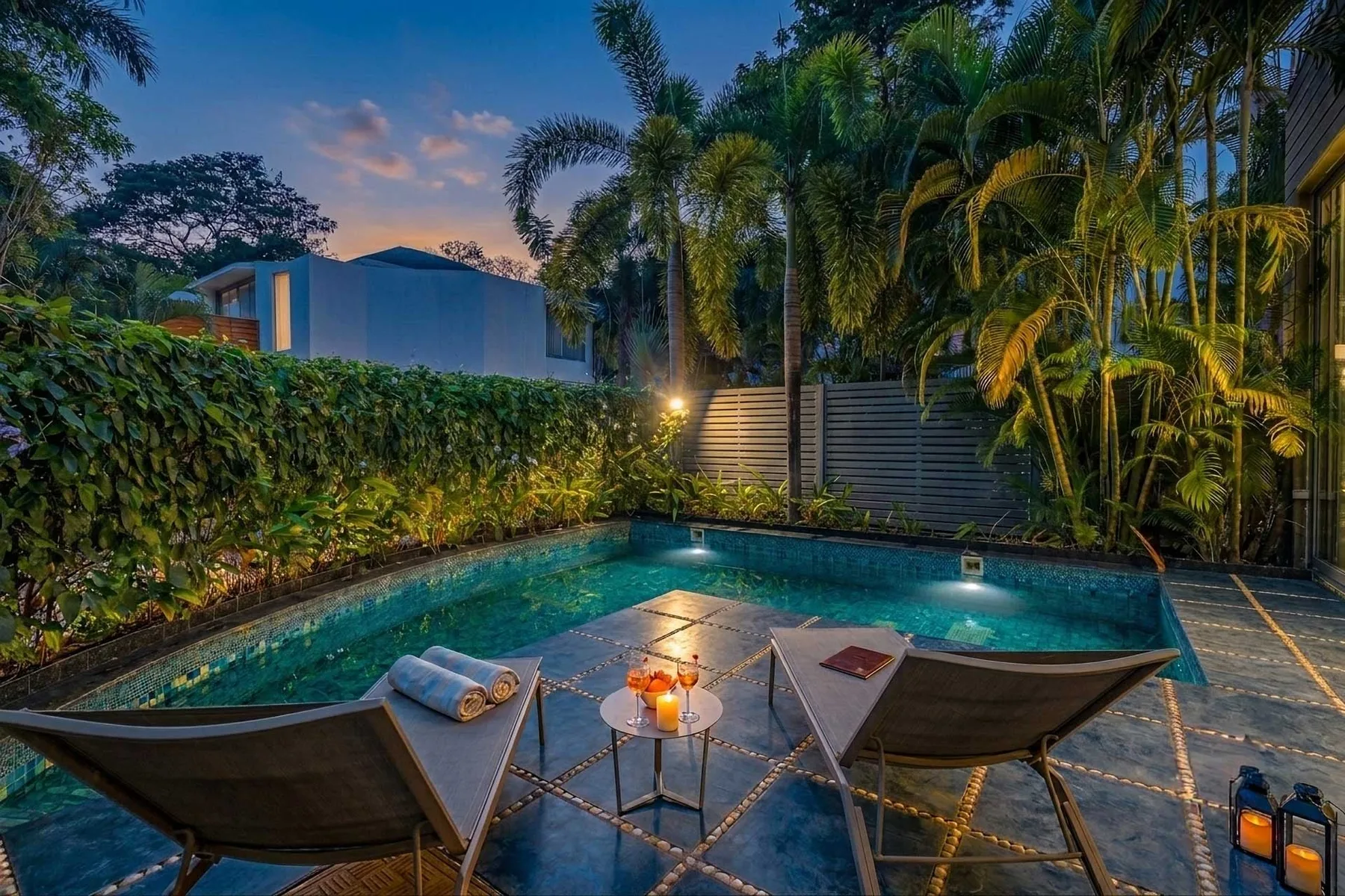 Una piscina exterior con asientos de descanso está rodeada de plantas tropicales al anochecer.
