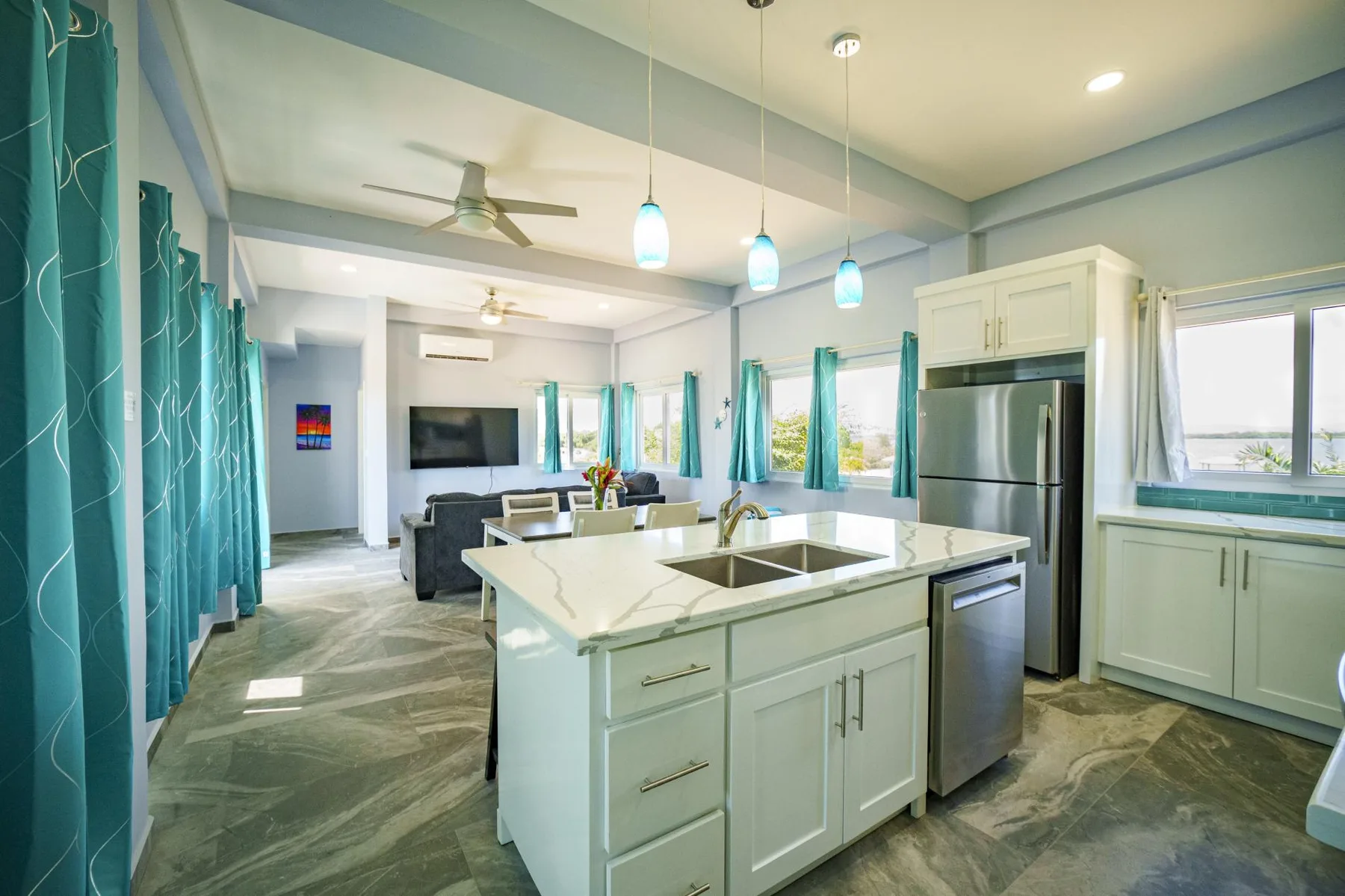 A bright open kitchen with a central island, stainless steel appliances, and teal curtains.