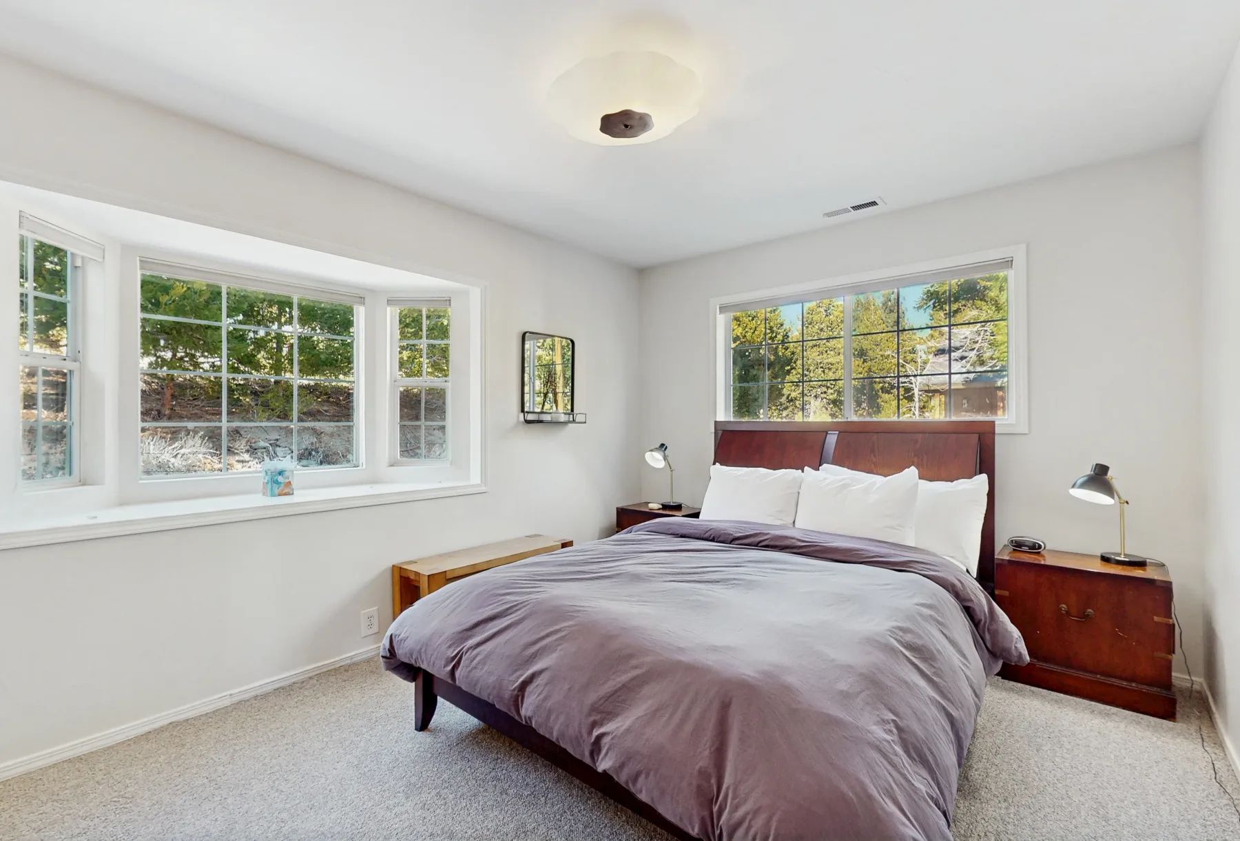 Un dormitorio con cama, mesitas de noche y ventanas con vista a los árboles.