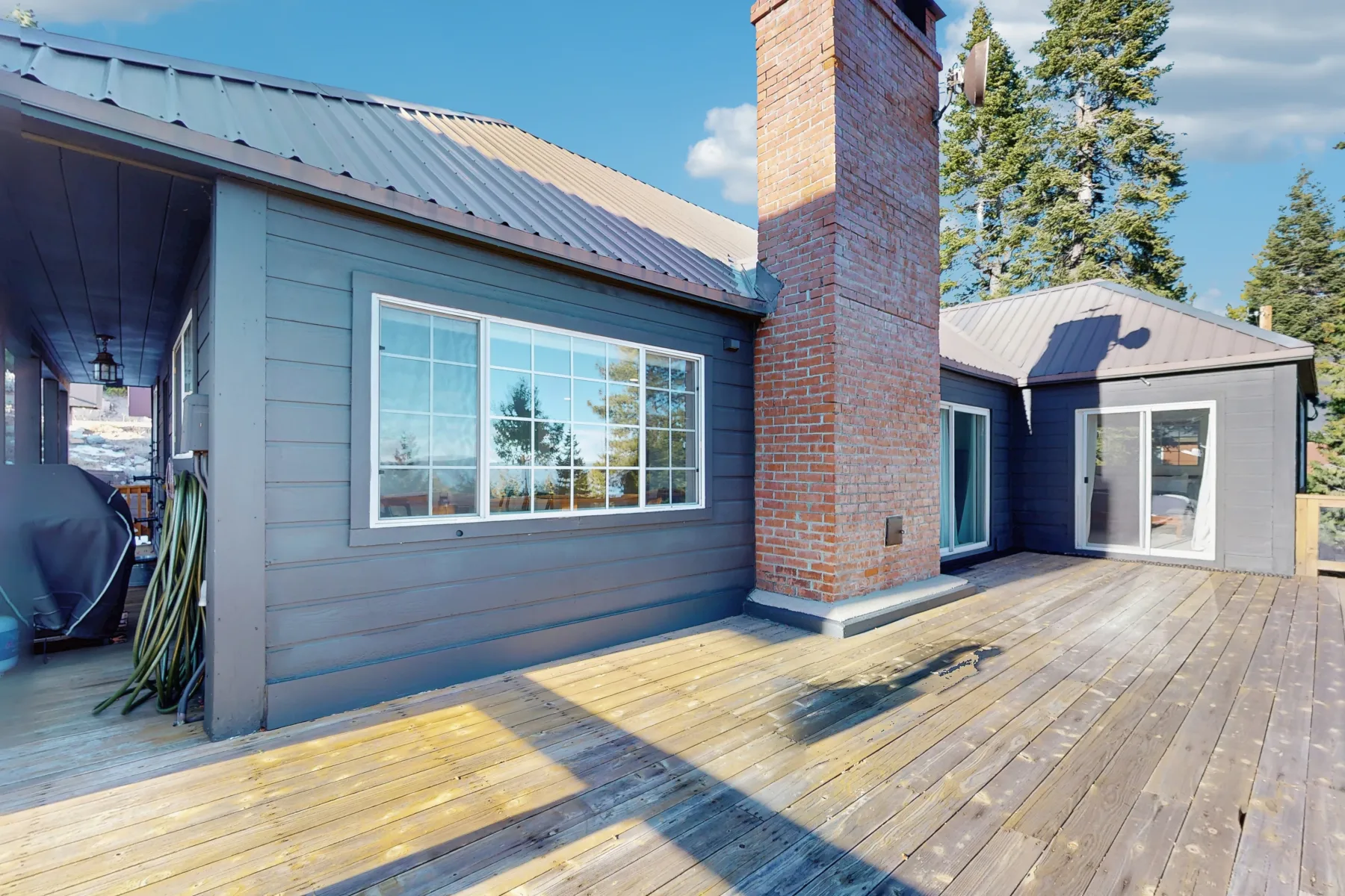 Una terraza de madera bordea una casa gris azulada con ventanas grandes y una chimenea de ladrillo.