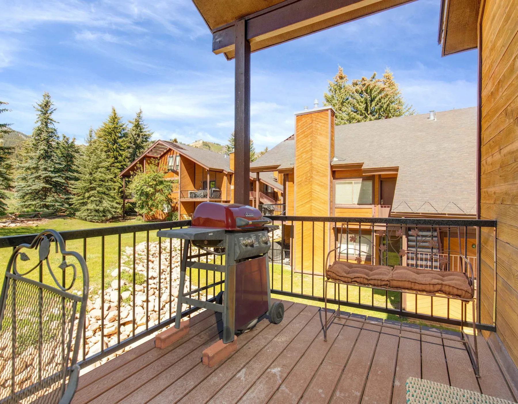 Una terraza de madera con una parrilla y un banco acolchado da a casas vecinas y árboles.