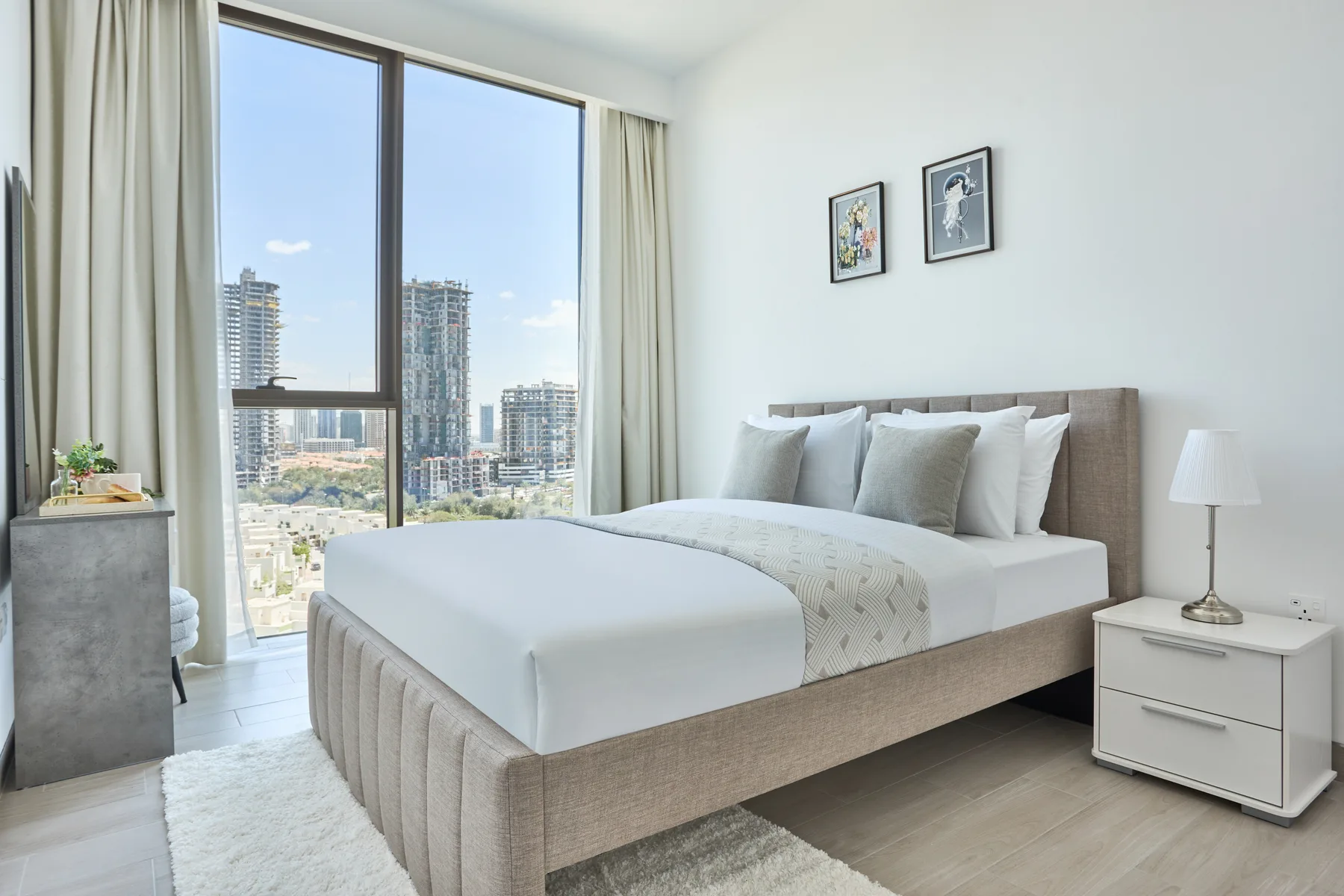 A bright bedroom features a bed beside large windows overlooking city buildings.