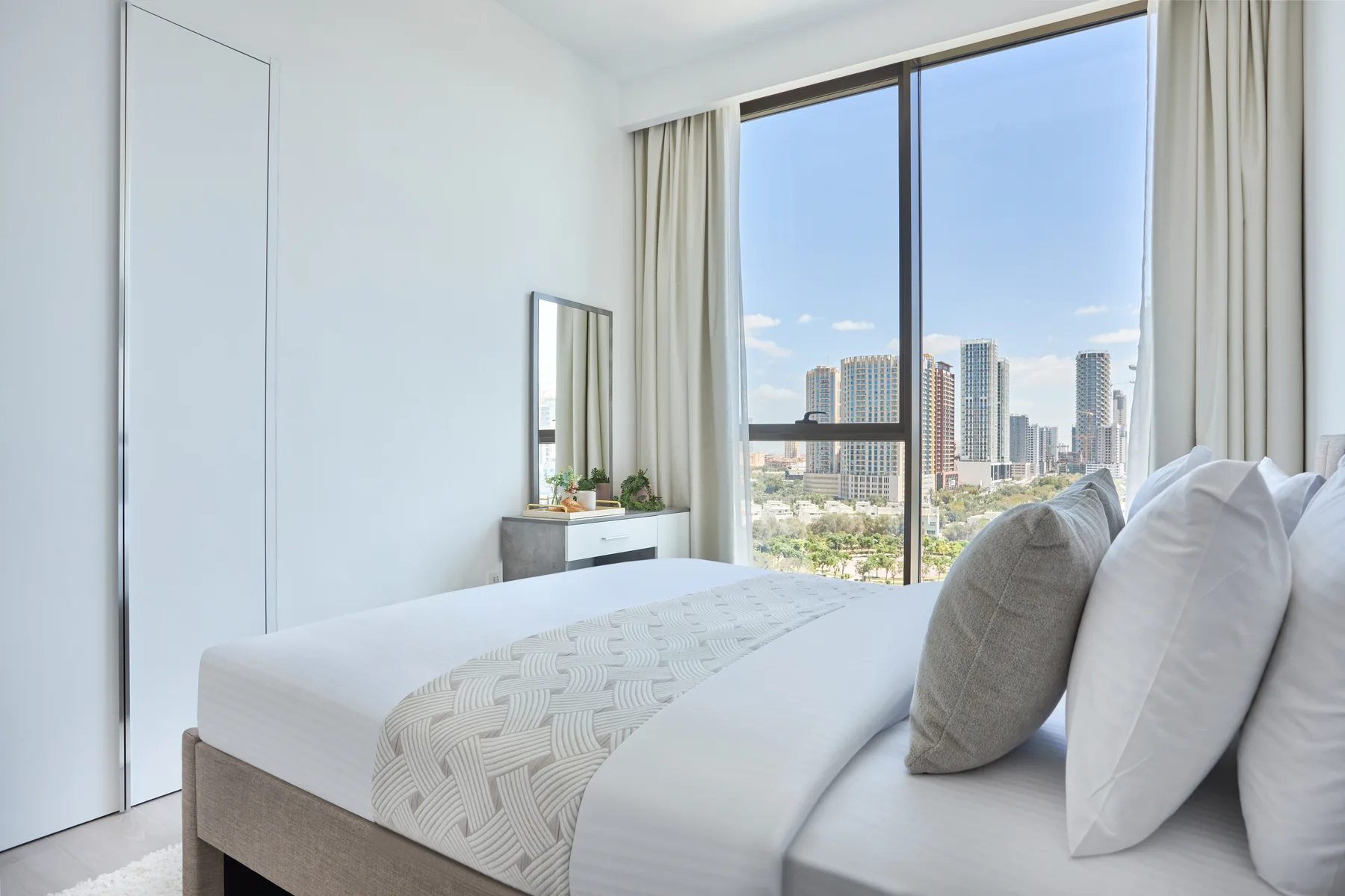 A bright bedroom with a neatly made bed beside a large city-view window.