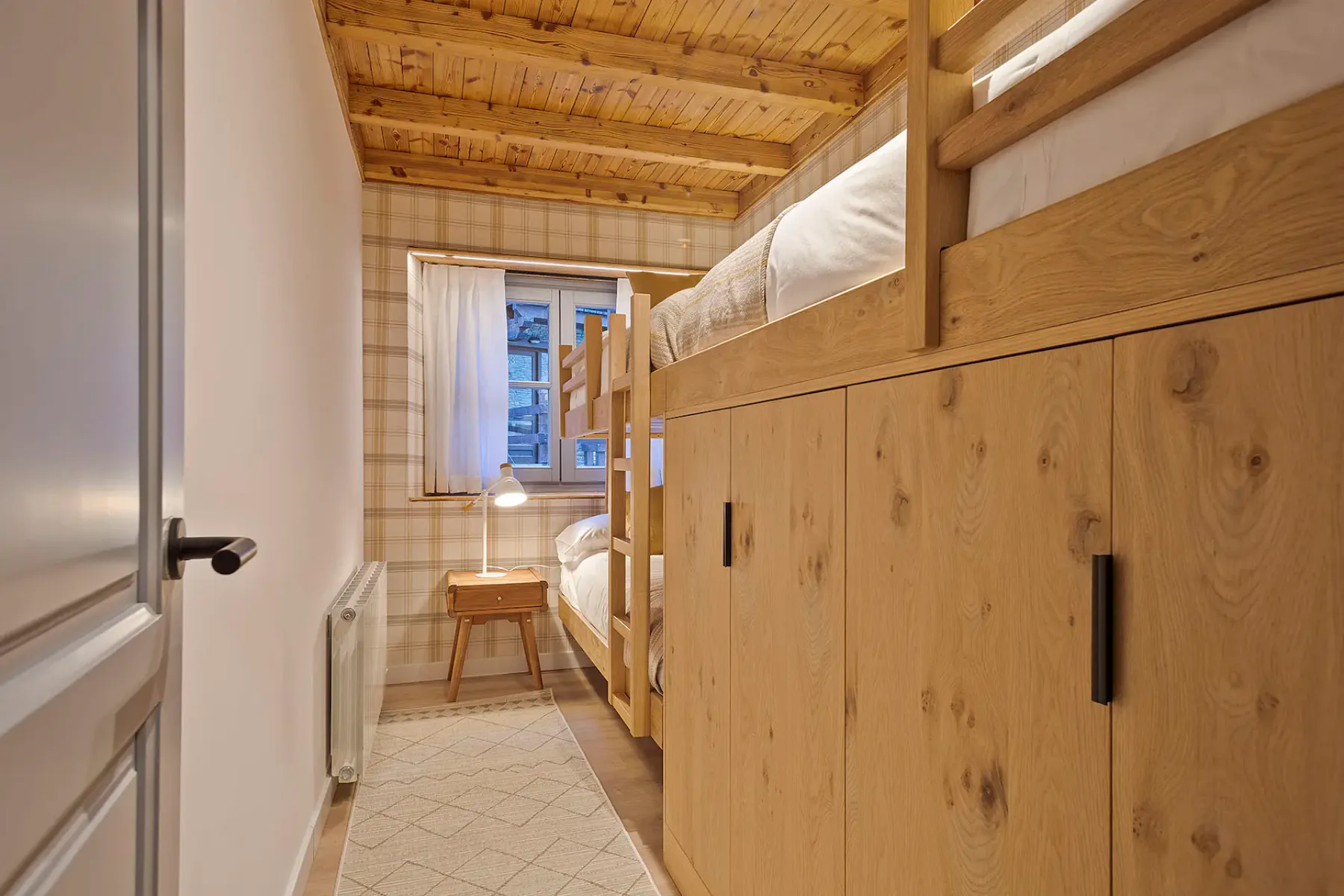 A compact bedroom with wooden bunk beds and a window at the far end.
