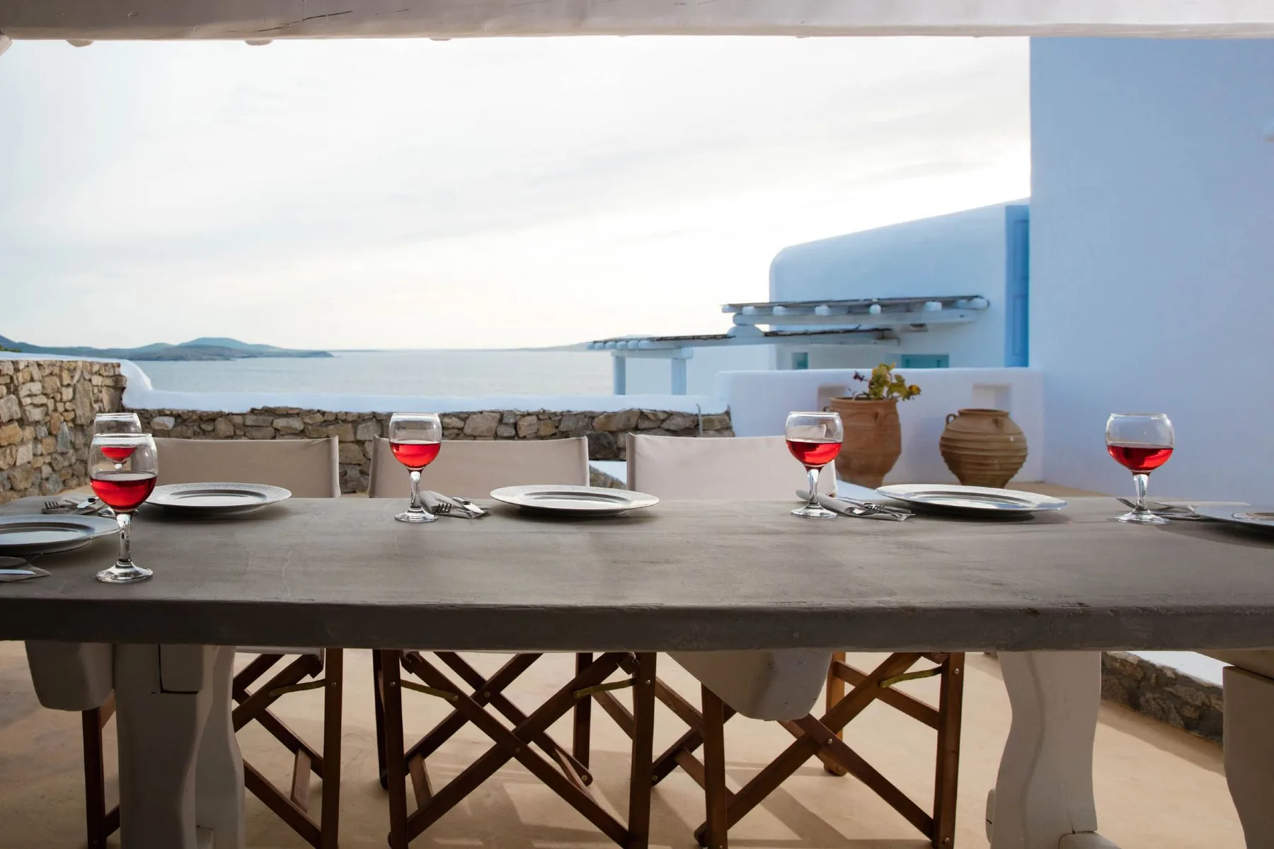 A dining table set with plates and red wine glasses overlooks the sea.