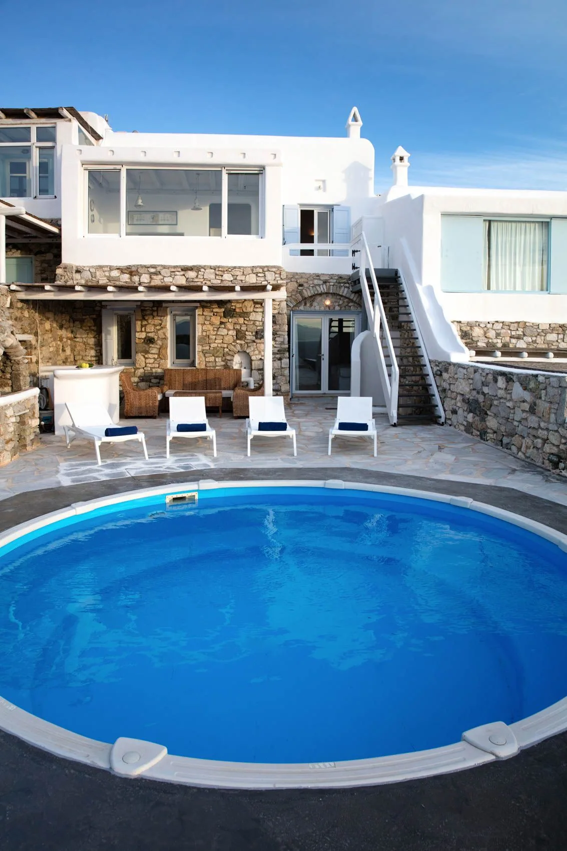 A swimming pool sits in front of a stone-and-white building with patio seating.