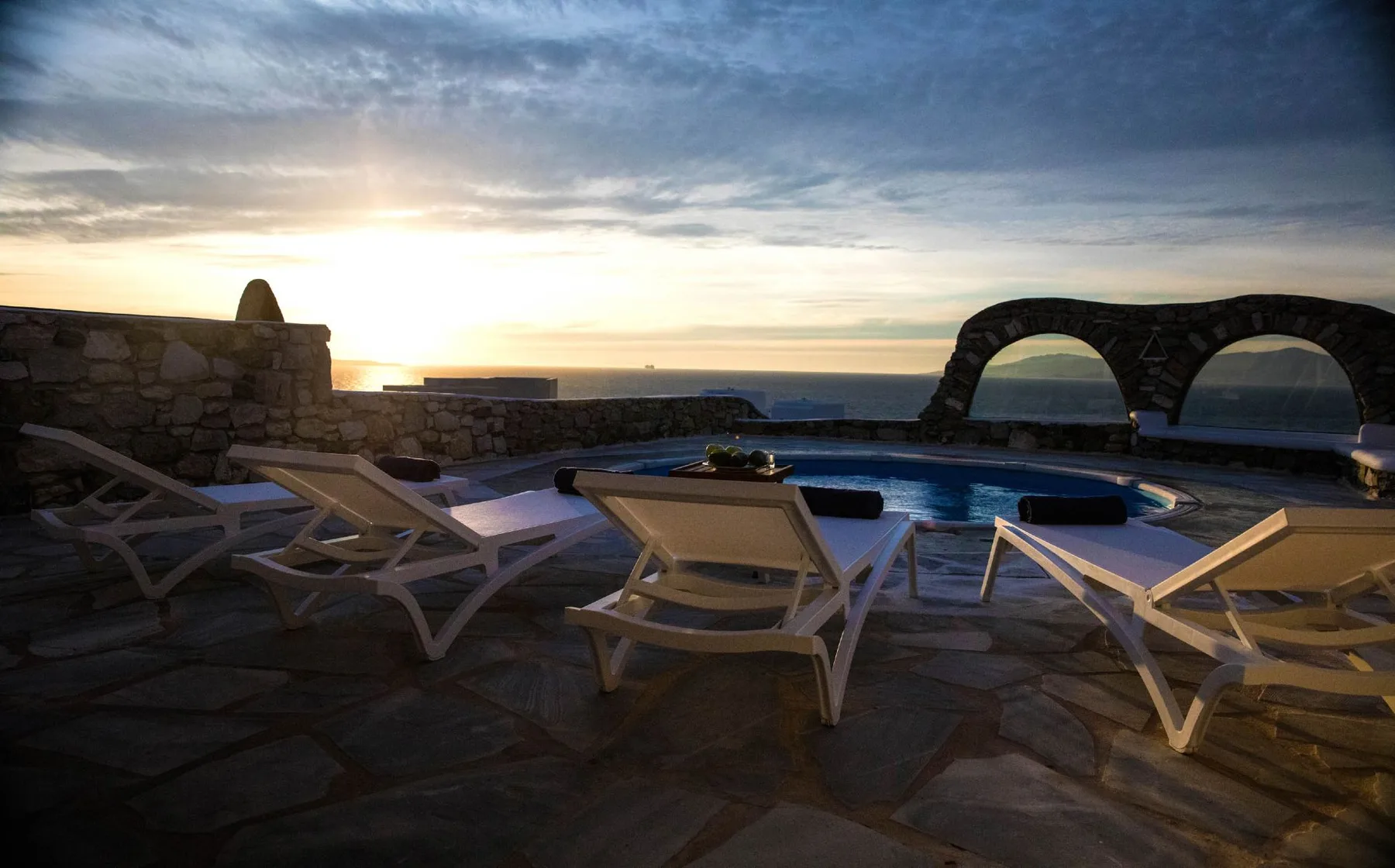 A stone pool terrace with lounge seating overlooks the sea at sunset.