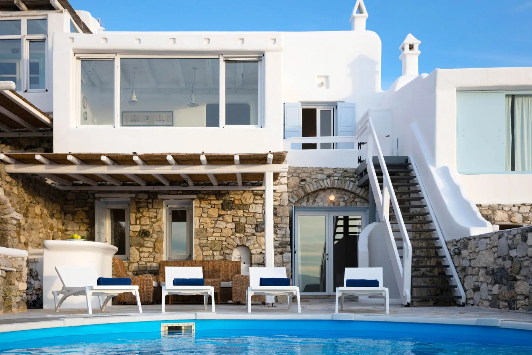A swimming pool sits in front of a white building with stone walls and an exterior staircase.