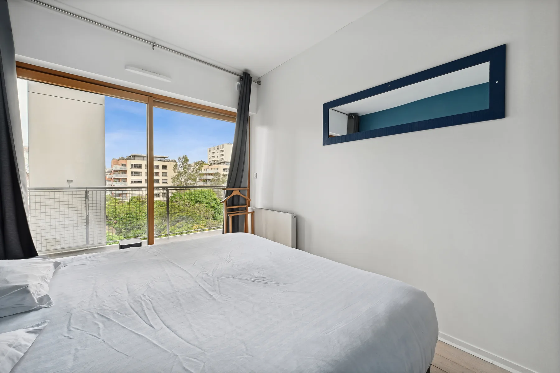 A bedroom with a bed beside large windows overlooking nearby buildings and greenery.