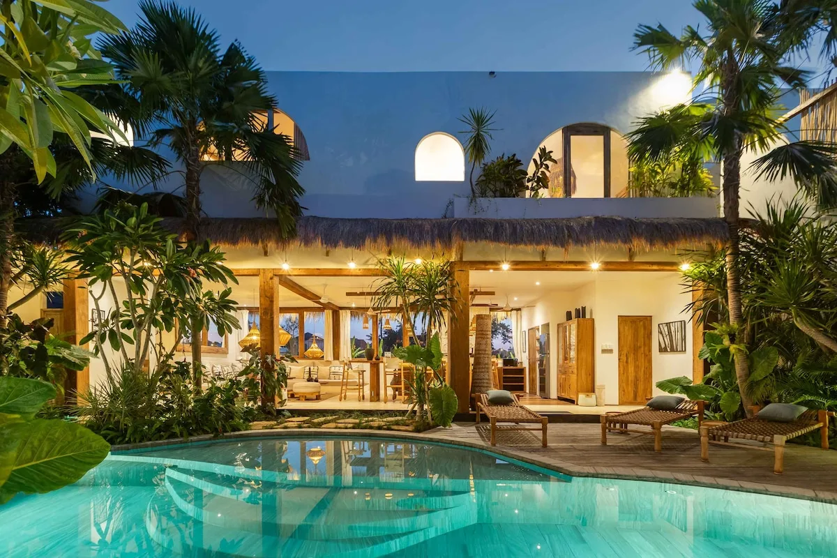 A tropical house opens onto a pool, with palms and outdoor seating.