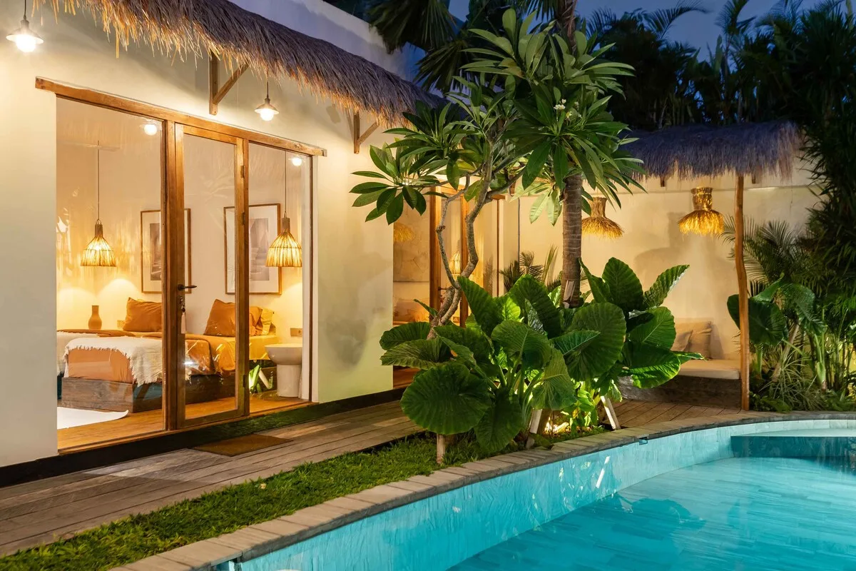 A poolside bungalow with a lit bedroom visible through glass doors.