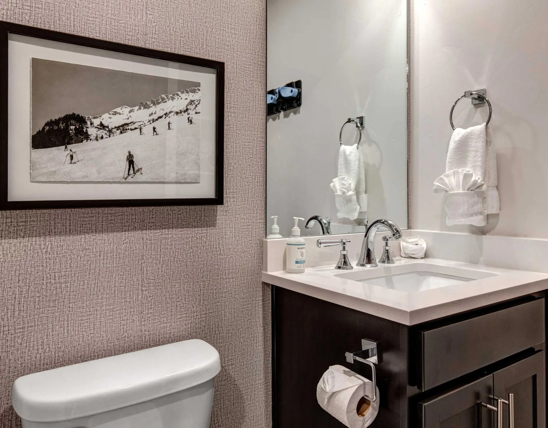 Un lavabo de baño con espejo está junto a una fotografía enmarcada de un paisaje nevado.
