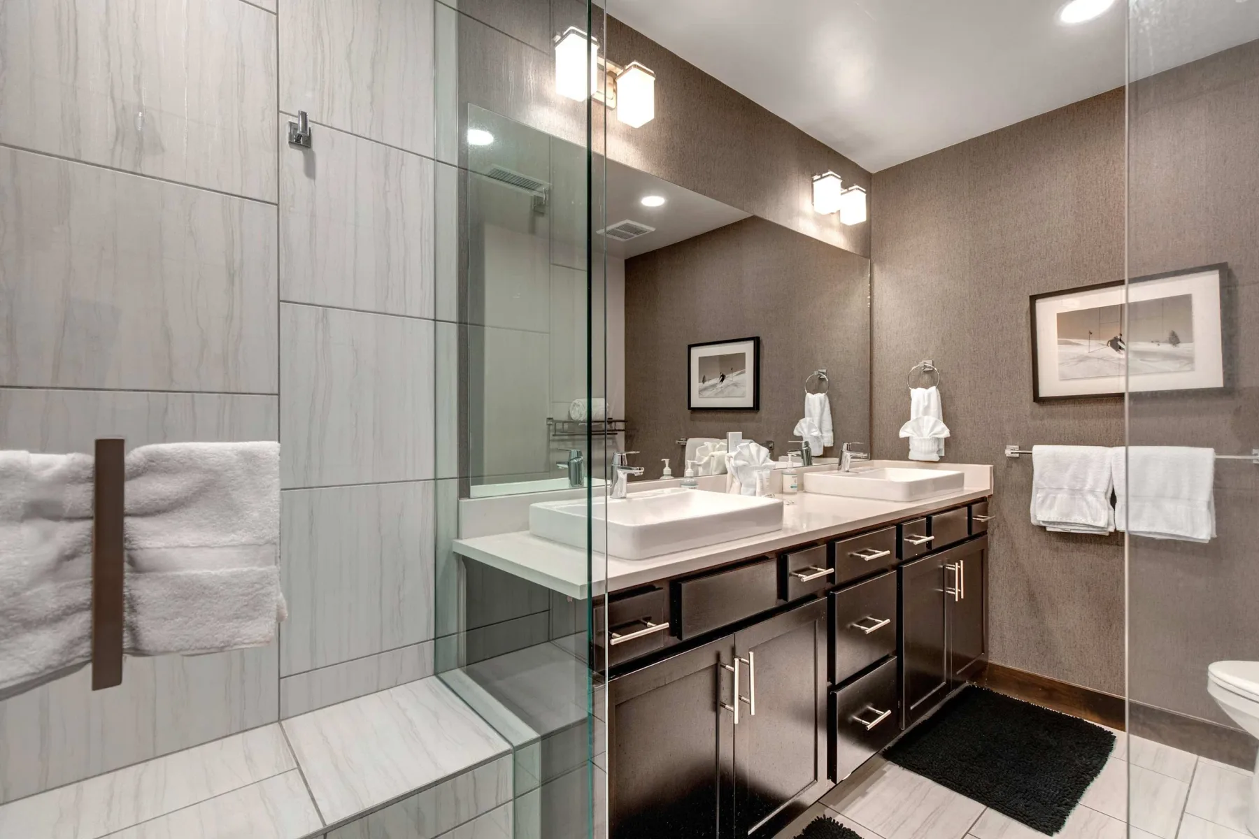 Un baño revestido de azulejos con ducha de vidrio y un largo tocador con dos lavabos.