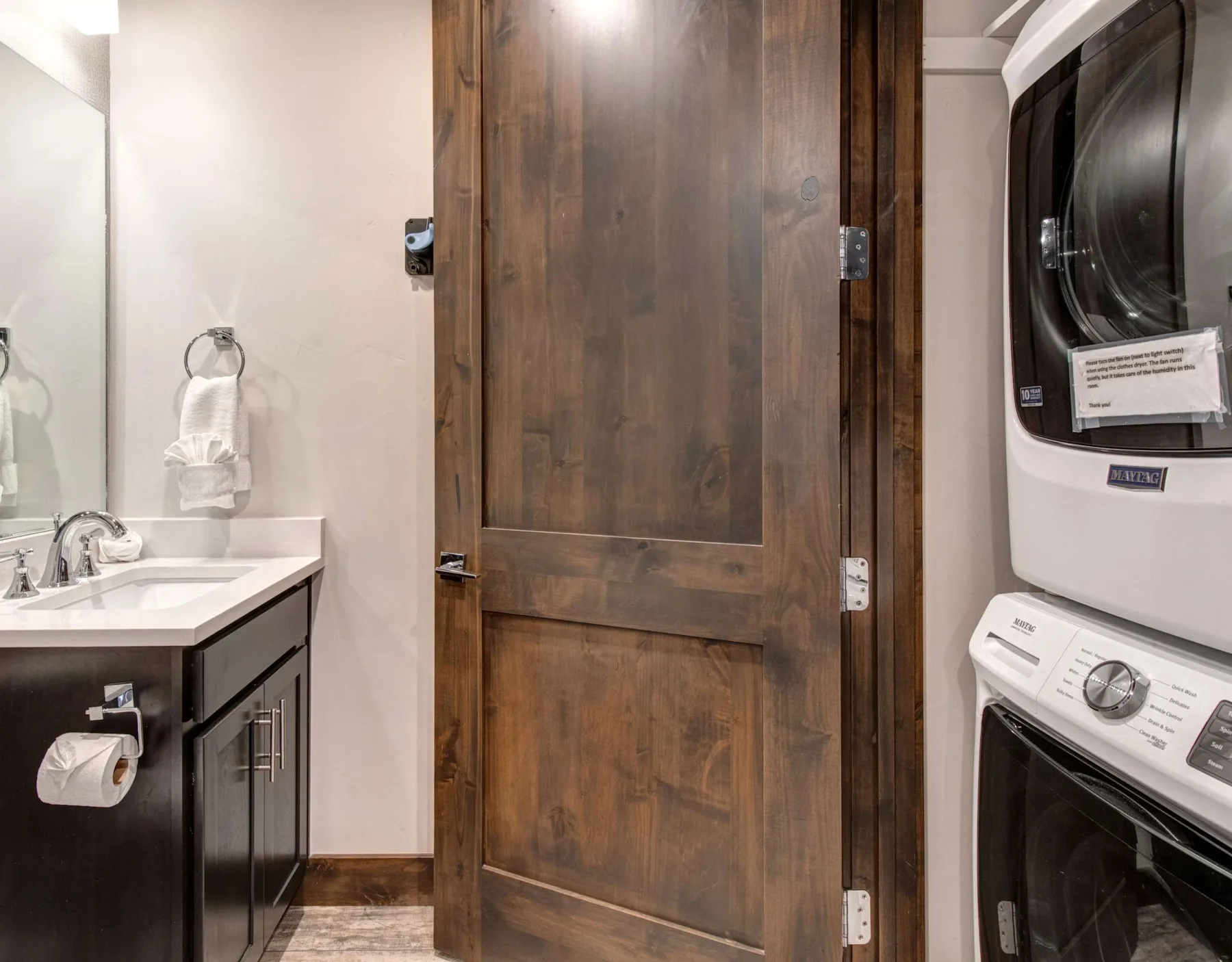 Un lavabo con espejo está junto a una puerta de madera y una lavadora y secadora apiladas.