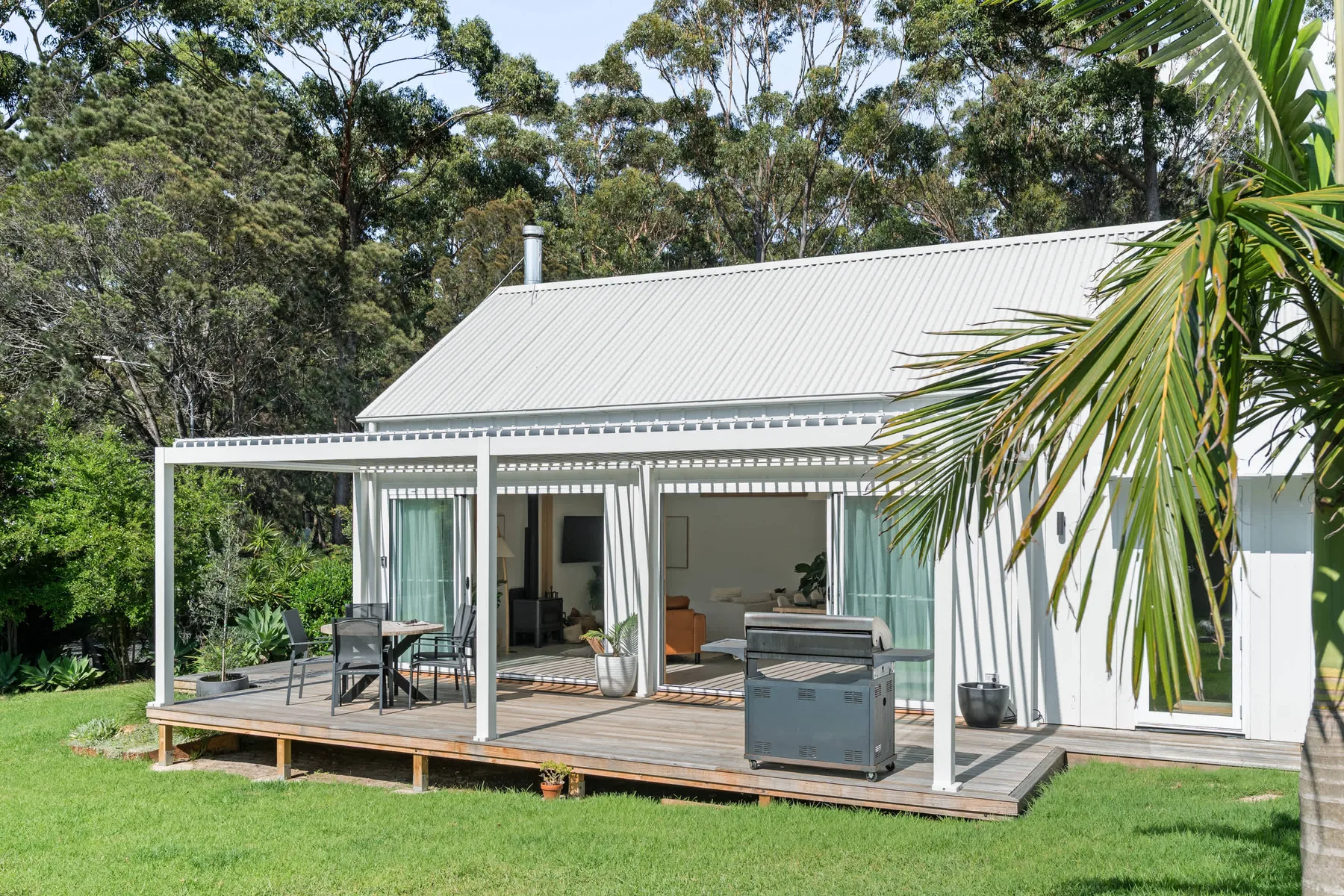 A white house with a covered wooden deck, outdoor seating, and a green lawn.