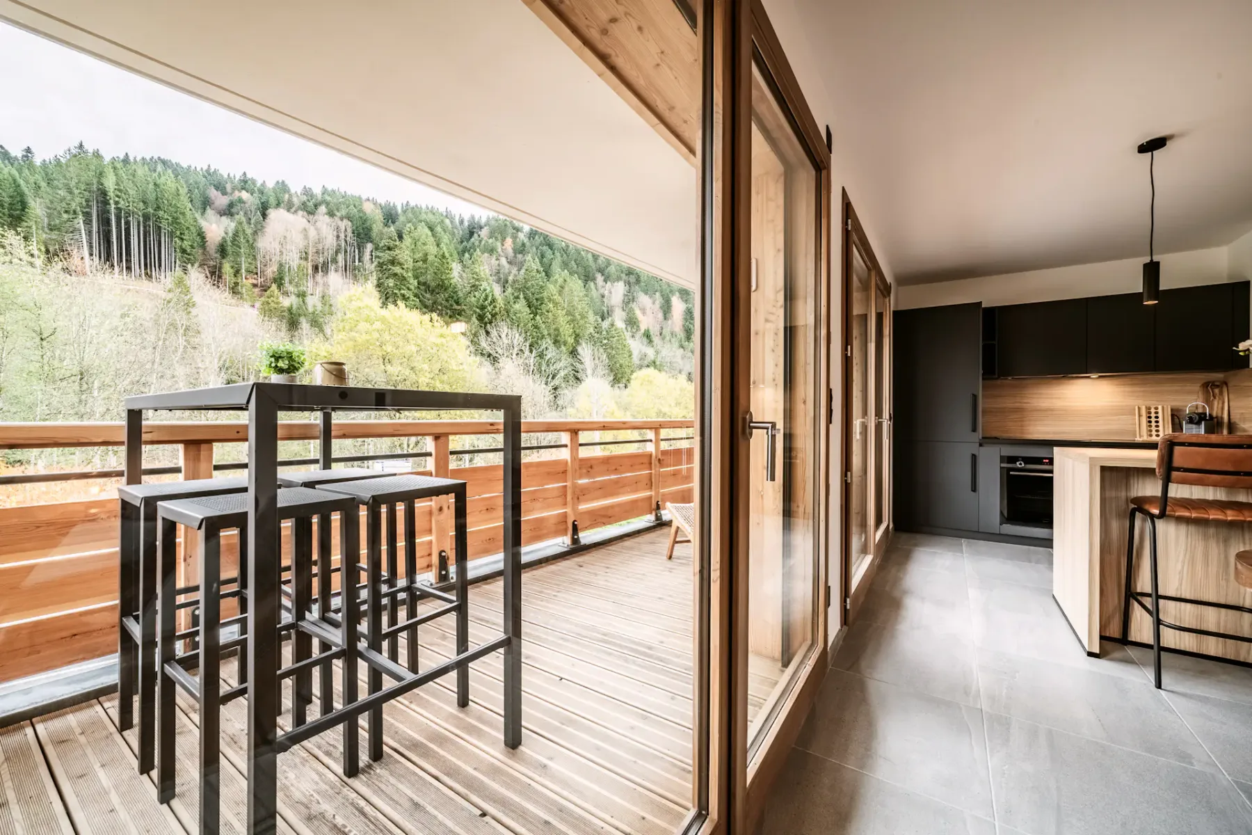 Una cocina se abre a una terraza de madera con asientos altos y vistas a una ladera boscosa.