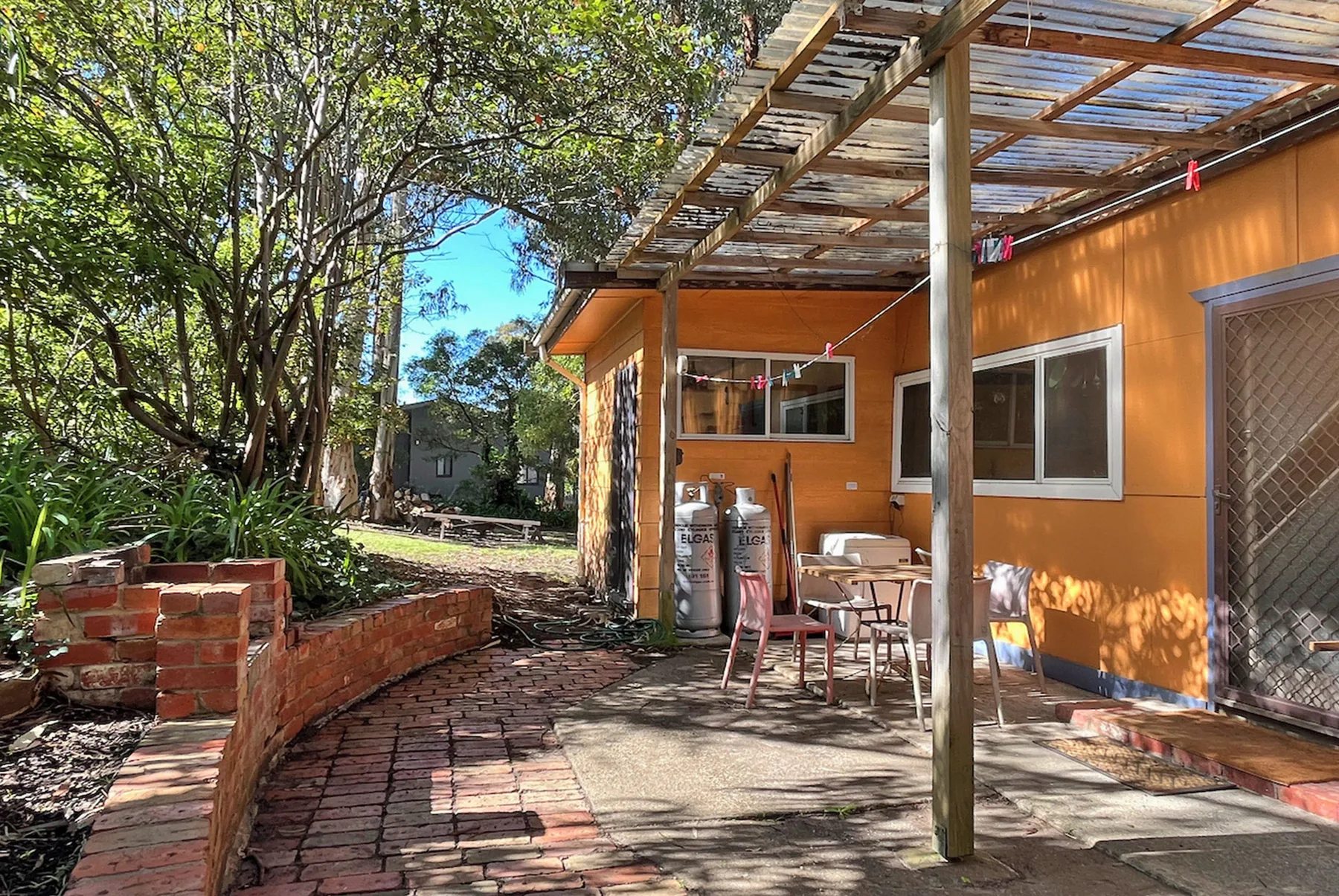 Un patio exterior junto a un edificio naranja, con asientos bajo una pérgola de madera.
