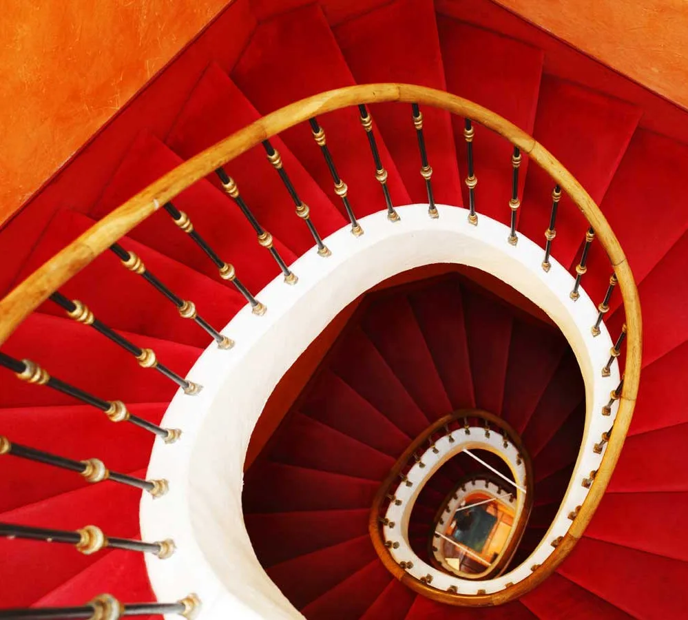 Representative hotel space: A spiral staircase curves downward with vivid red steps and a pale railing.