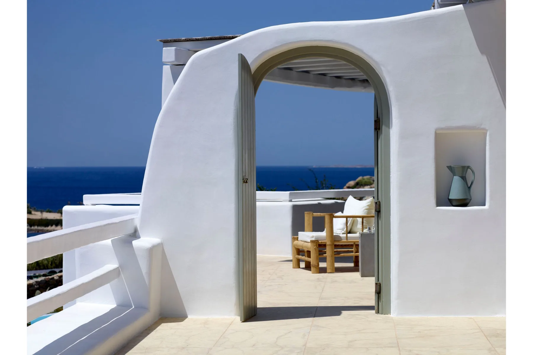 Una terraza blanca se abre por una puerta arqueada hacia asientos con vistas al mar.