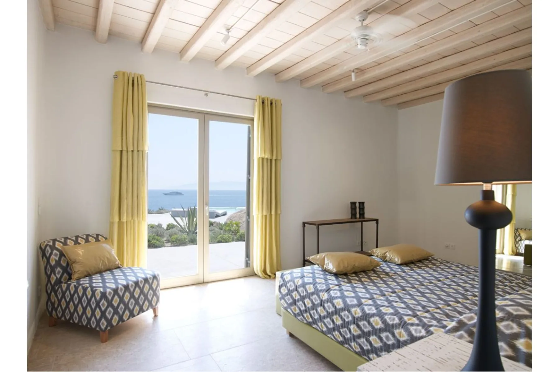 Un dormitorio con una cama estampada y asientos junto a puertas de cristal con vistas al mar.