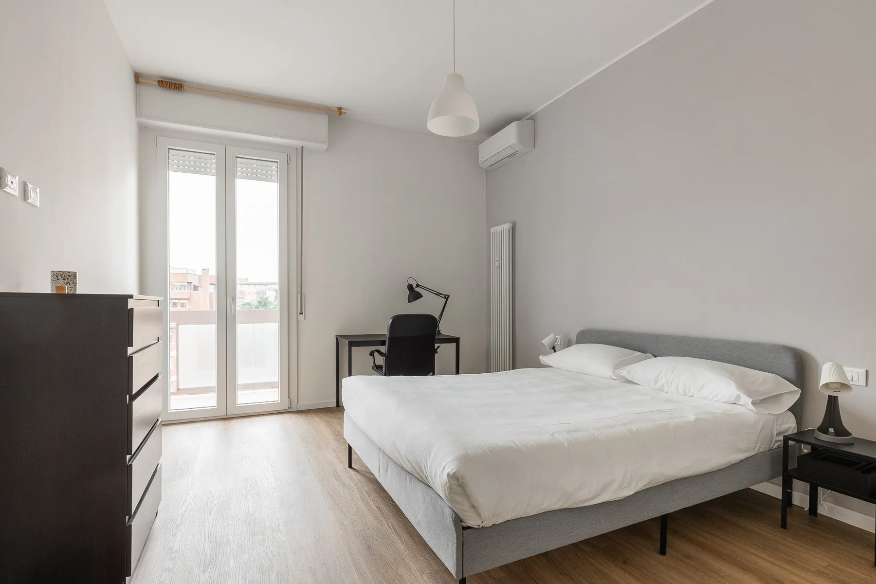 Un dormitorio luminoso con cama, escritorio y puertas de vidrio que dan a un balcón.