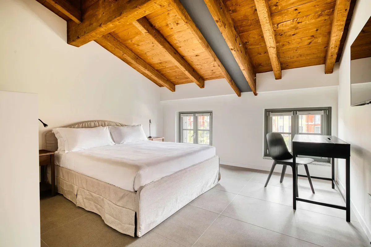 A bedroom with a large bed and a desk beneath exposed wooden ceiling beams.