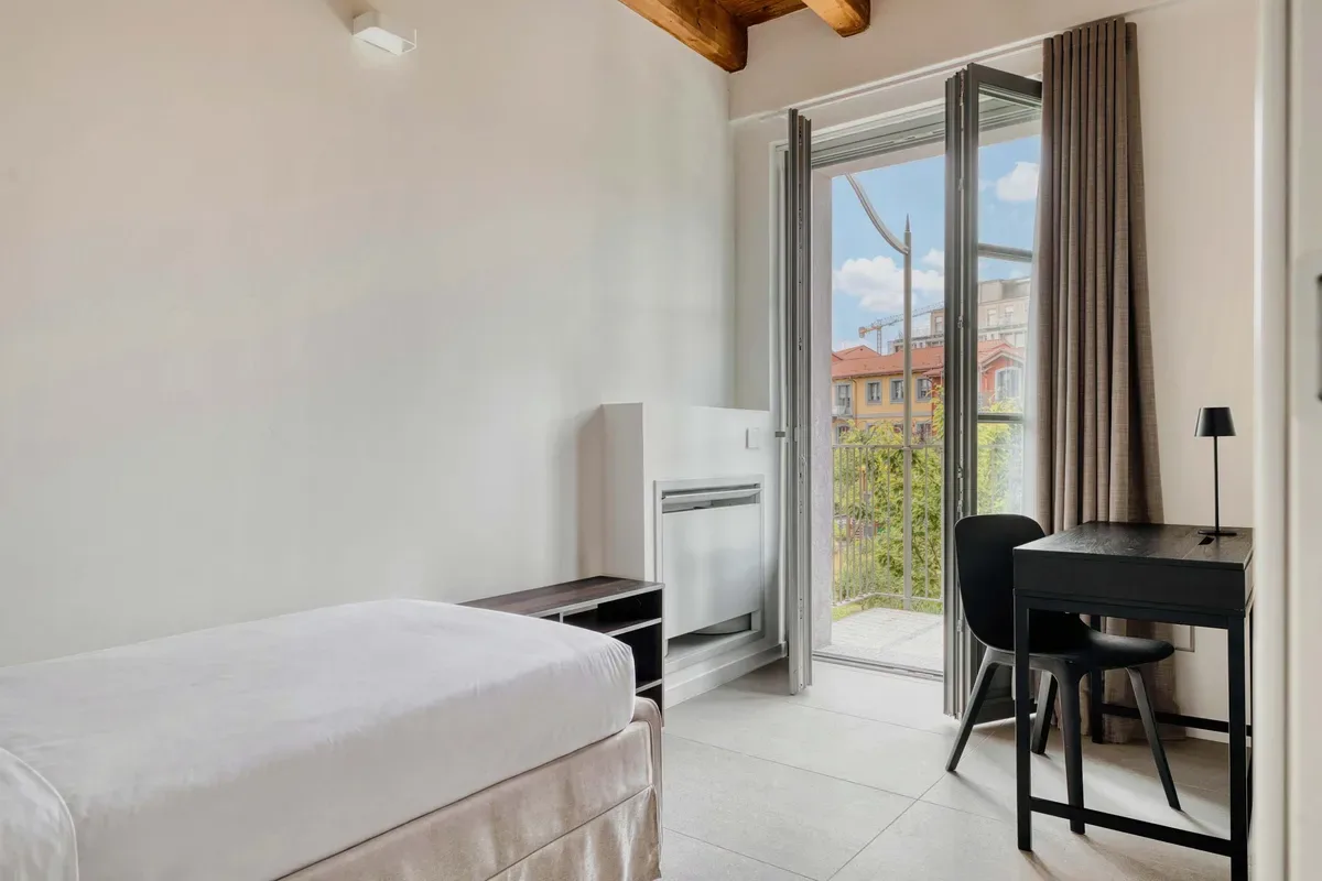 A bright bedroom with a bed, desk, and glass doors opening to a balcony.