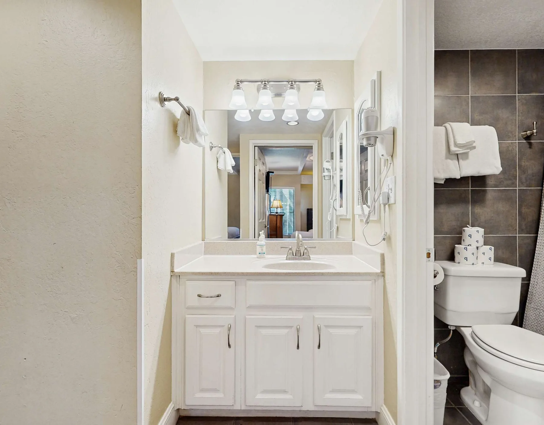 Un tocador de baño con lavabo y espejo, junto a una zona de inodoro con azulejos.