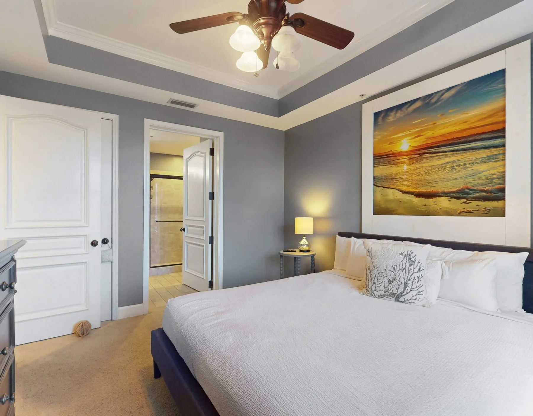 A bedroom with a large bed, bedside lamp, and framed ocean sunset artwork.