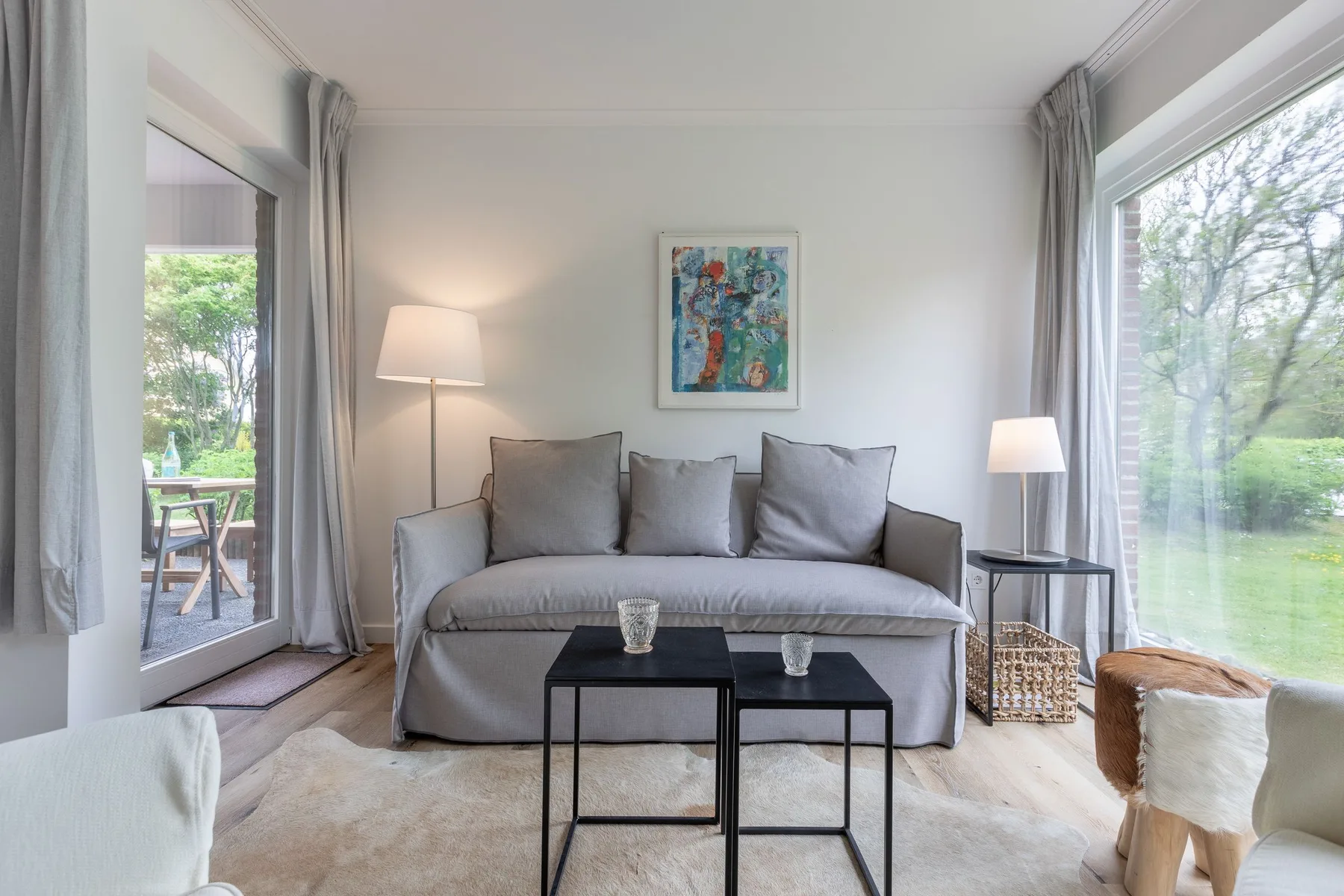 A bright living room with a gray sofa and large windows overlooking greenery.