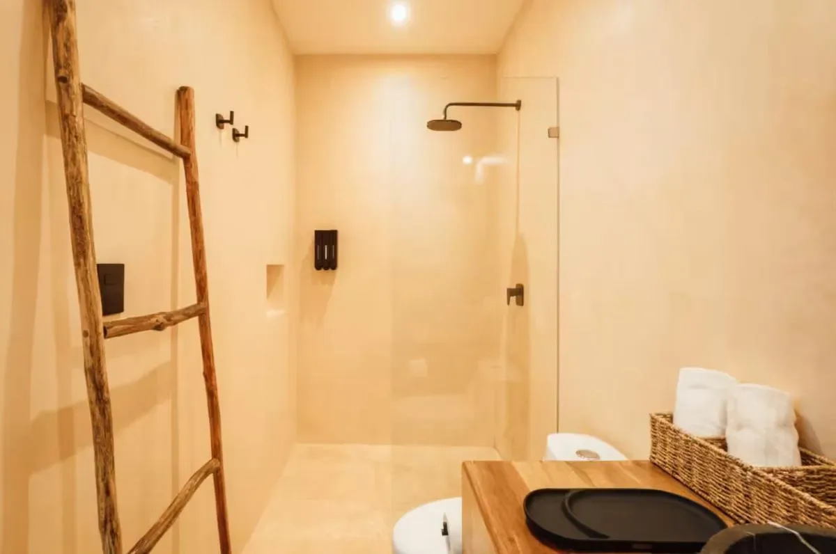 A warm-toned bathroom features a glass shower, wooden ladder towel rack, and a wooden vanity with a woven basket.