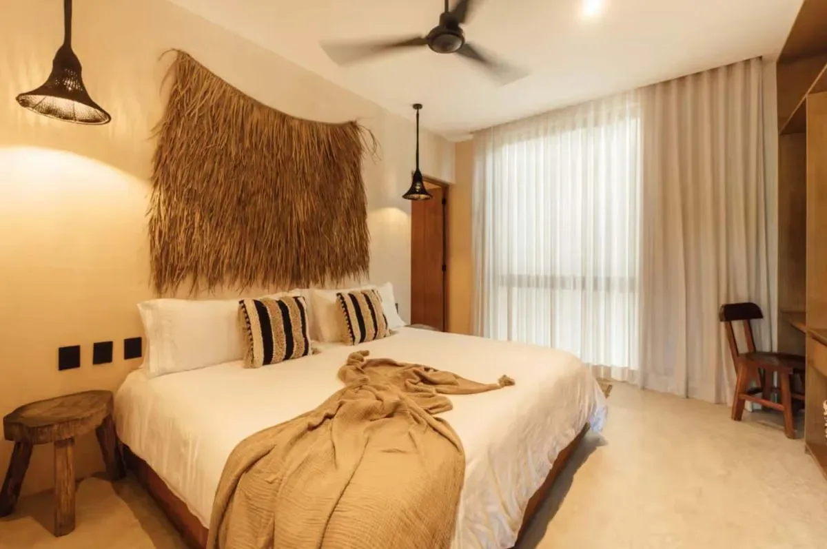 A bedroom with a white bed, woven wall hanging, striped pillows, and sheer curtains.