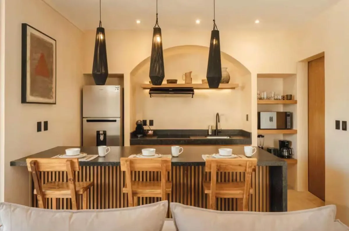 A warm-toned kitchen features a central island with wooden stools, pendant lights, and built-in shelving.