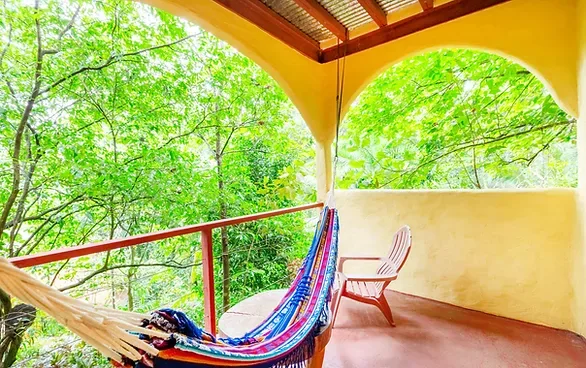 Representative hotel space: A covered balcony with a hammock and chair overlooks leafy trees.