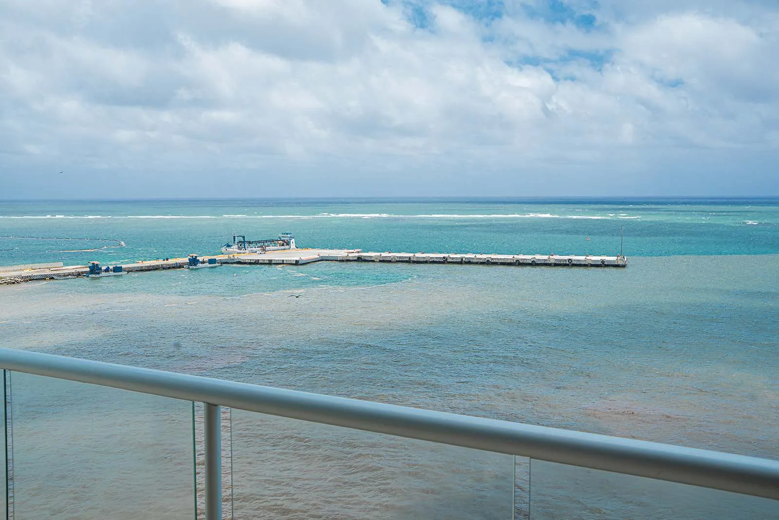 Vista costera desde un balcón, con aguas turquesas tranquilas y un muelle bajo.